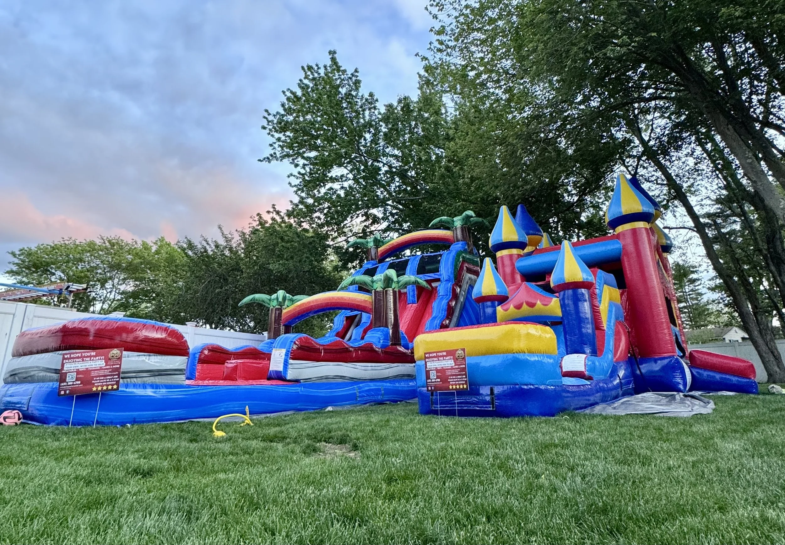 Colorful inflatable water slide with castles, palm trees, and rainbow designs set up on a grassy backyard, with green trees and blue sky in the background.