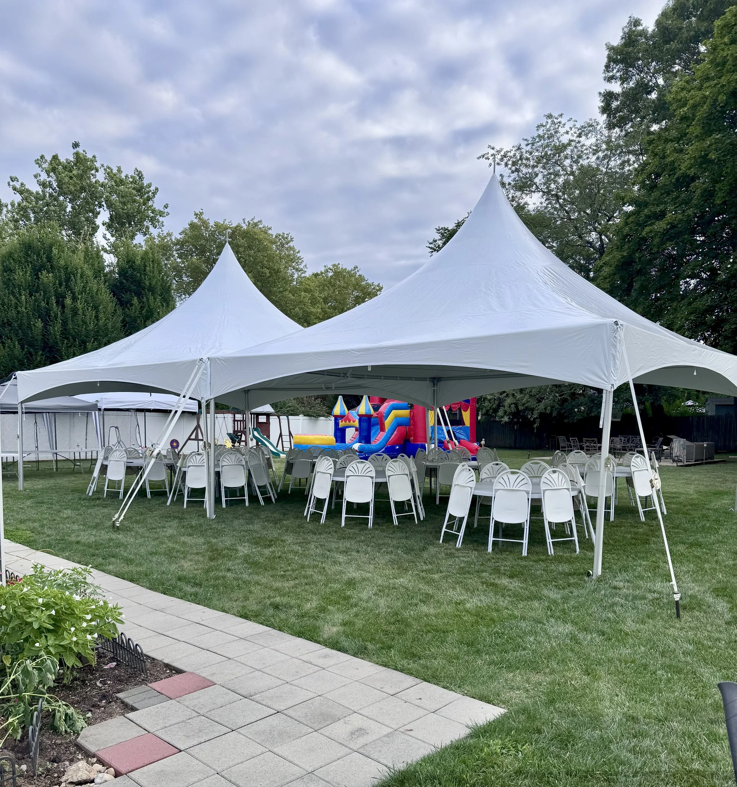 White event tents set up in a backyard with folding chairs and tables underneath, rainbow-colored inflatable bounce house and slide in the background, green lawn, trees, cloudy sky.