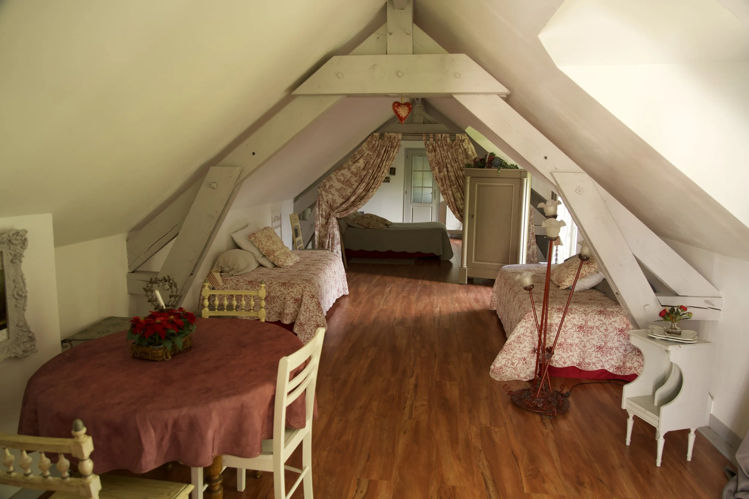 Grenier aménagé avec plafond en pente, deux lits simples, une table avec nappe, et un lit double à l'arrière. Décoration rustique et chaleureuse.