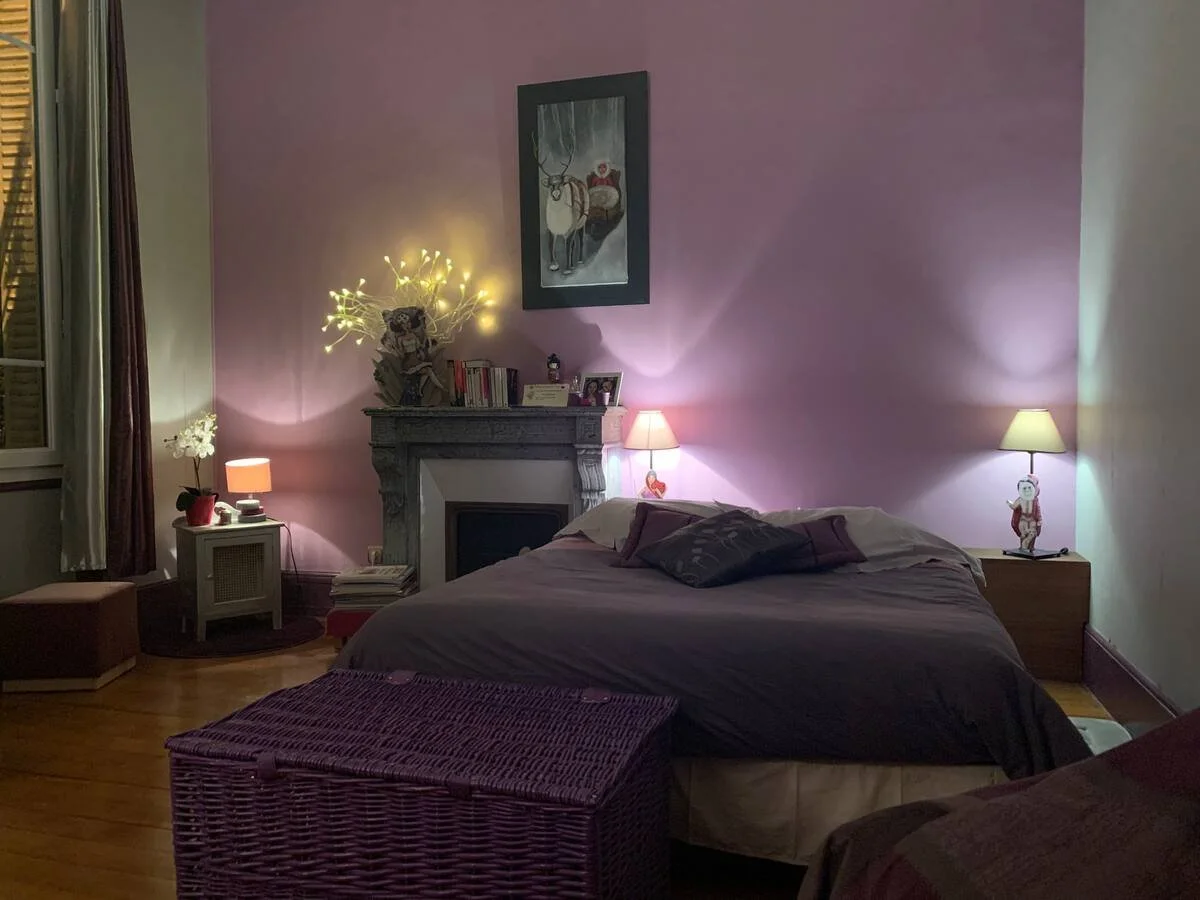 Chambre à coucher avec mur violet, lit avec couverture grise, lampes de chevet allumées, cheminée décorative, tableau et panier en osier violet.