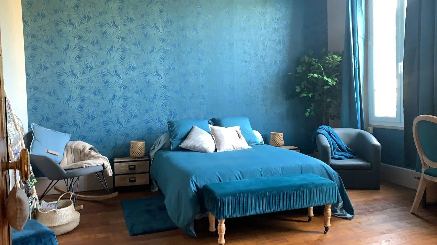 Chambre avec mur bleu, lit avec couvre-lit bleu, coussins blancs, table de chevet, fauteuil, rideaux et plante dans un coin.