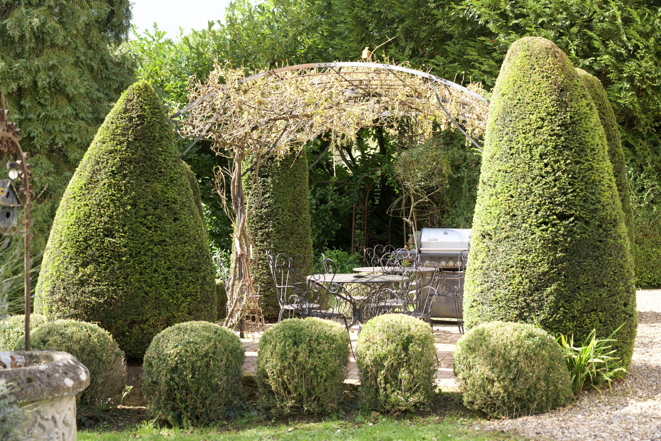 Une terrasse de jardin avec des buissons taillés, une tonnelle en fer forgé, des meubles de jardin en métal et un barbecue en acier inoxydable.