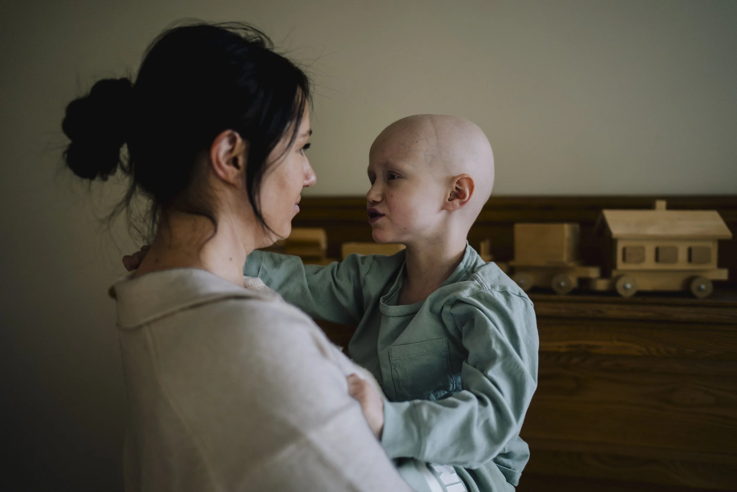 A woman holding a young boy with a shaved head and visible scars, in a room with wooden furniture.