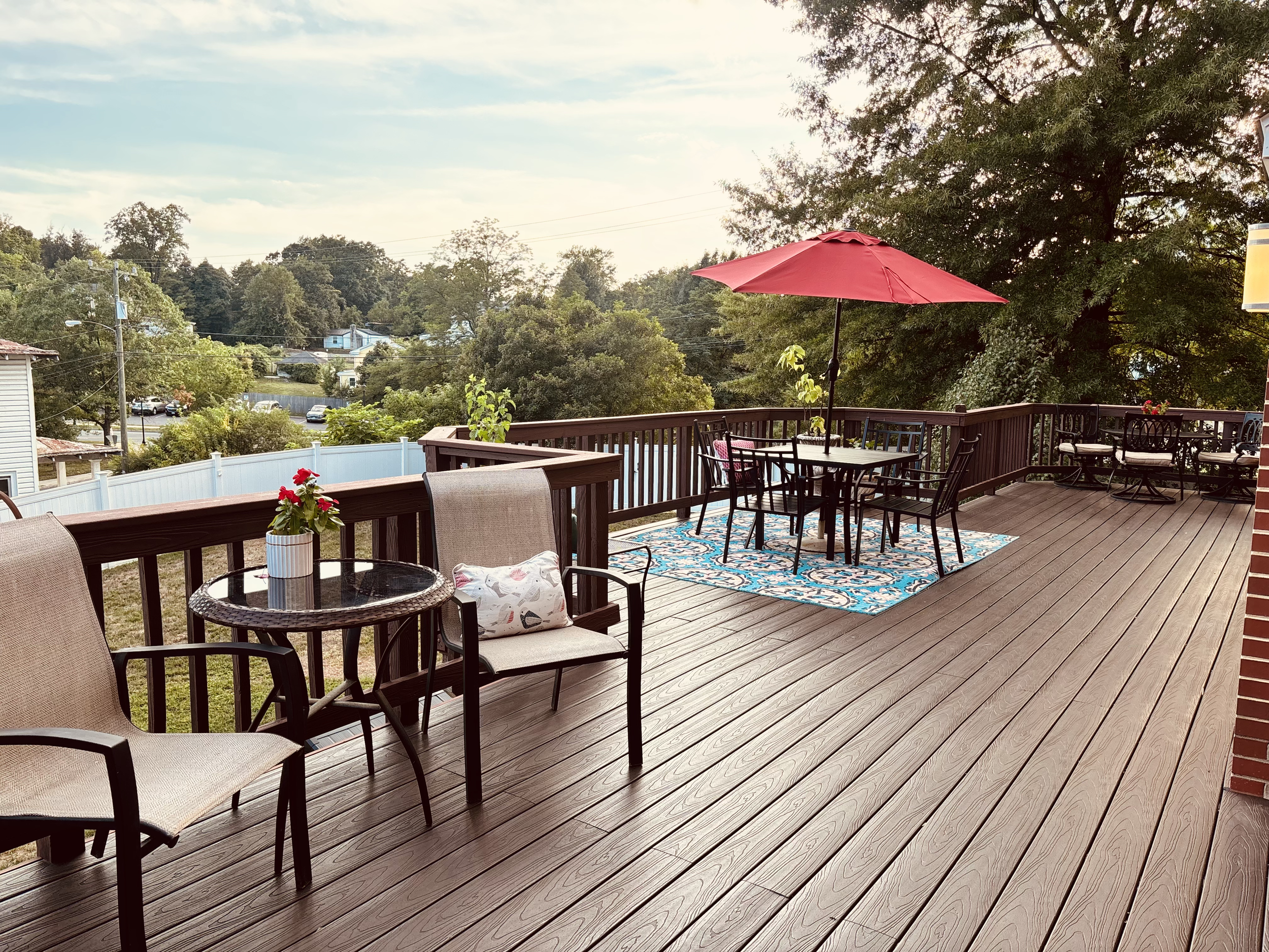 Spacious wooden deck attached to a red-brick building, with outdoor furniture and trees in the background.