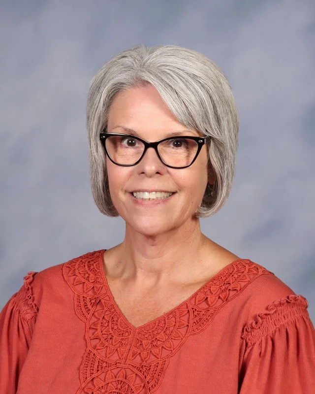 A smiling elderly woman with gray hair, wearing glasses and a blue patterned blouse, standing outdoors in a park with trees in the background.