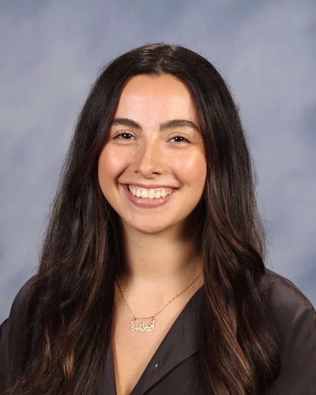 A young woman with long wavy hair wearing a red dress, smiling outdoors in a park with green trees in the background.