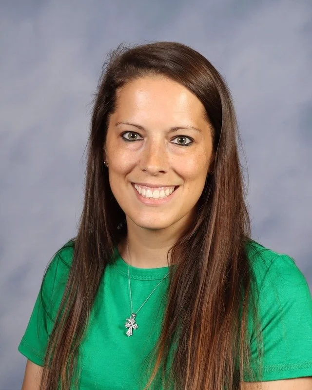 A smiling young woman with long brown hair wearing a red shirt standing outdoors in a park with green trees in the background.