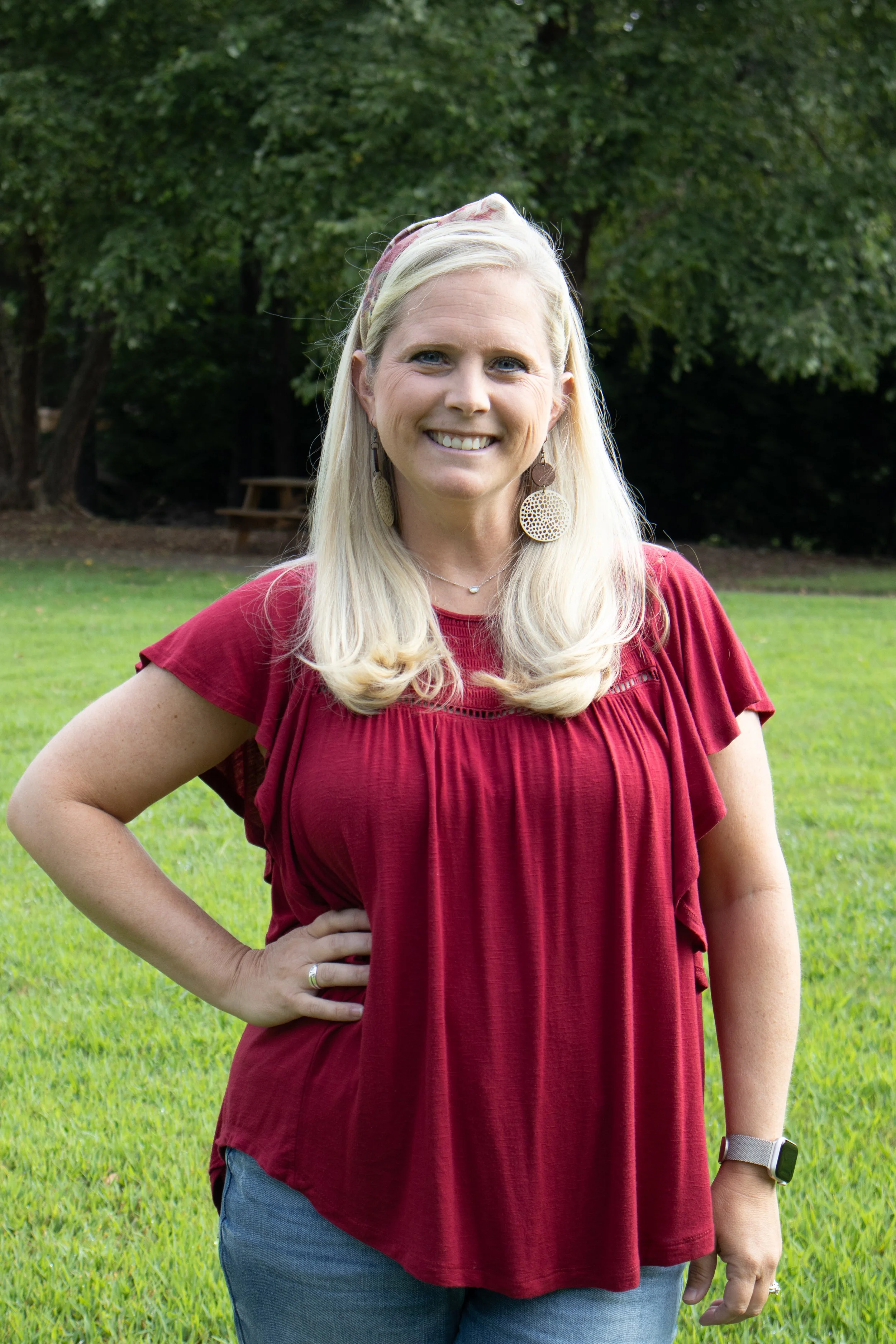 A woman with long blonde hair, wearing a red top, blue jeans, large earrings, and a watch, standing outdoors on grass with trees in the background.