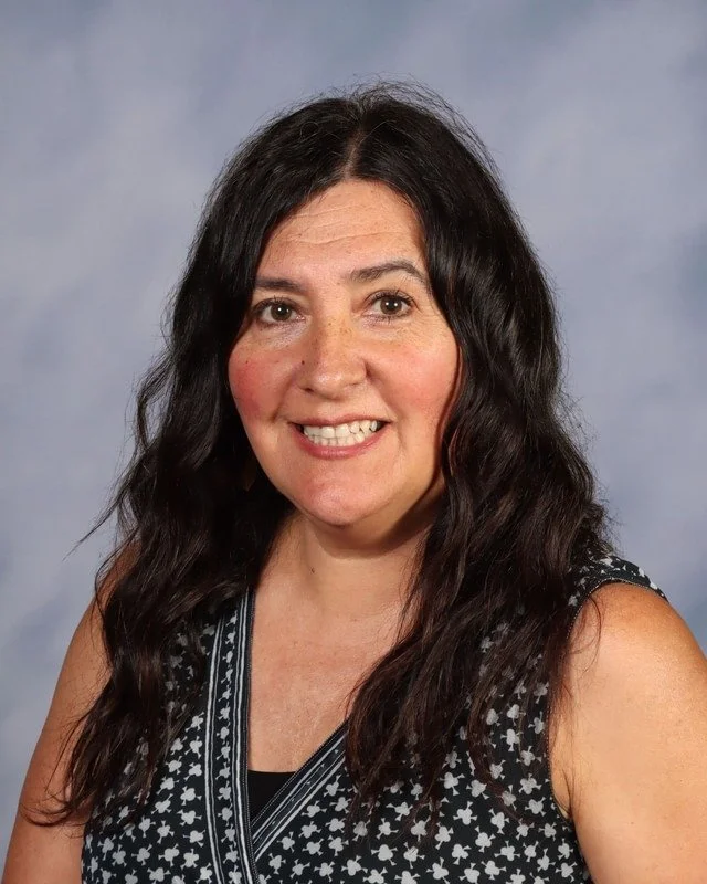A woman with long dark hair smiling outdoors on a grassy area with trees in the background.