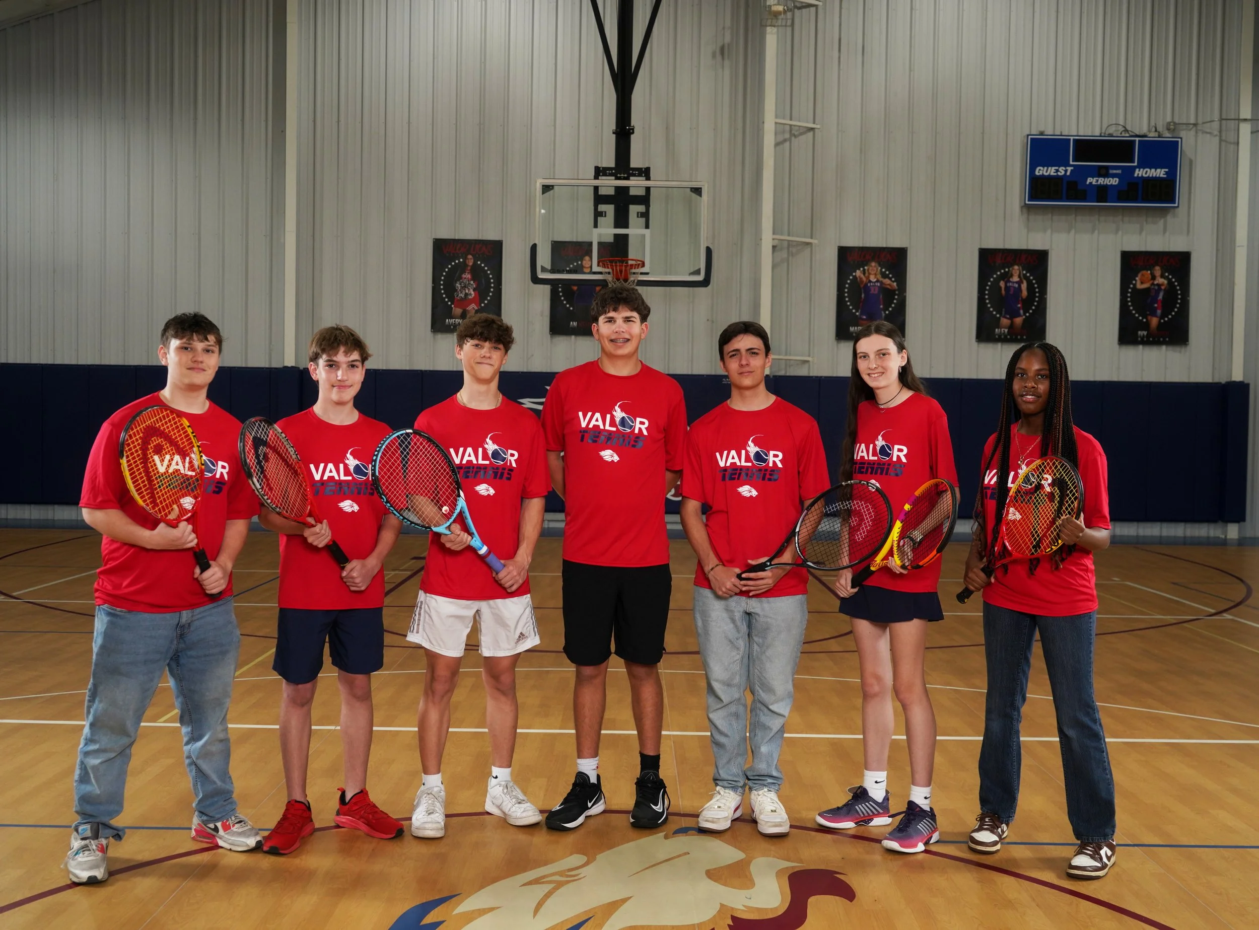 A group of seven young girls in red sports shirts and skirts standing in a gymnasium on a basketball court, holding tennis rackets.
