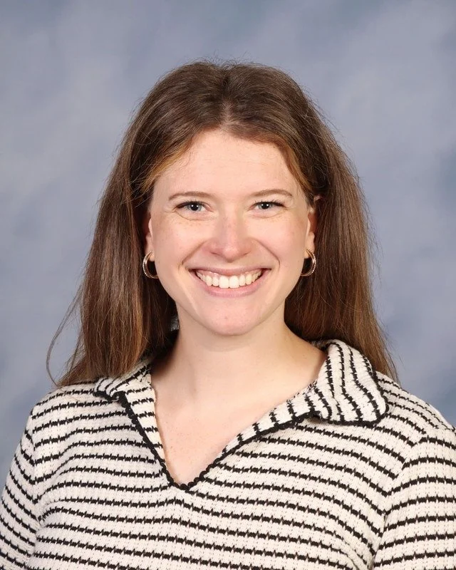 A smiling woman with brown hair, wearing a black and white floral sleeveless top, standing outdoors in a park with green trees in the background.