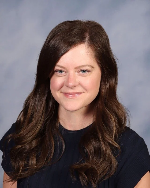 A woman with long brown hair wearing a navy blue sweater, standing outdoors on grass with trees in the background.