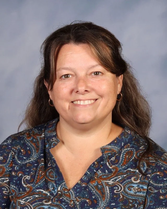 A woman with dark hair and bangs, wearing a navy blue top, standing outdoors with trees and greenery in the background, smiling at the camera.