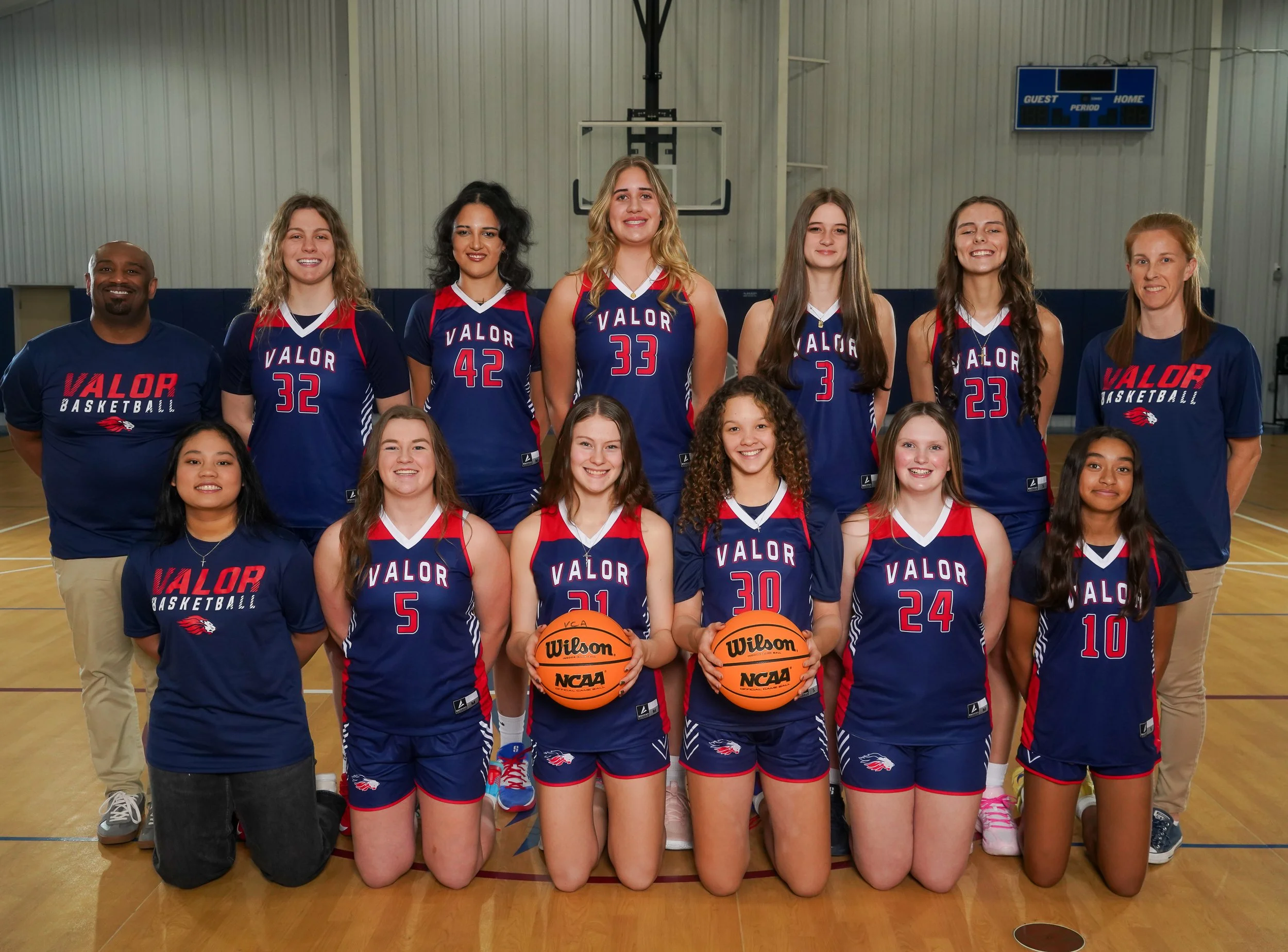 A women's volleyball team posing in a gymnasium, wearing navy blue uniforms with red accents, with some members kneeling in the front row and others standing behind, along with two coaches.
