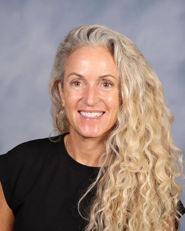 A smiling middle-aged woman with curly gray hair standing outdoors in a lush green park on a sunny day.
