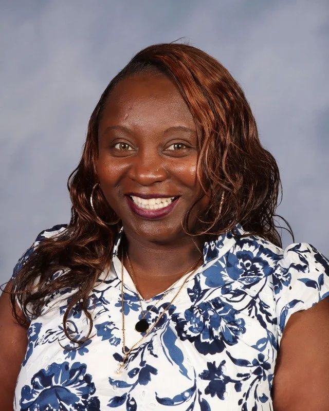 Smiling woman in an olive green jacket and orange shirt standing outdoors in a park with trees in the background.