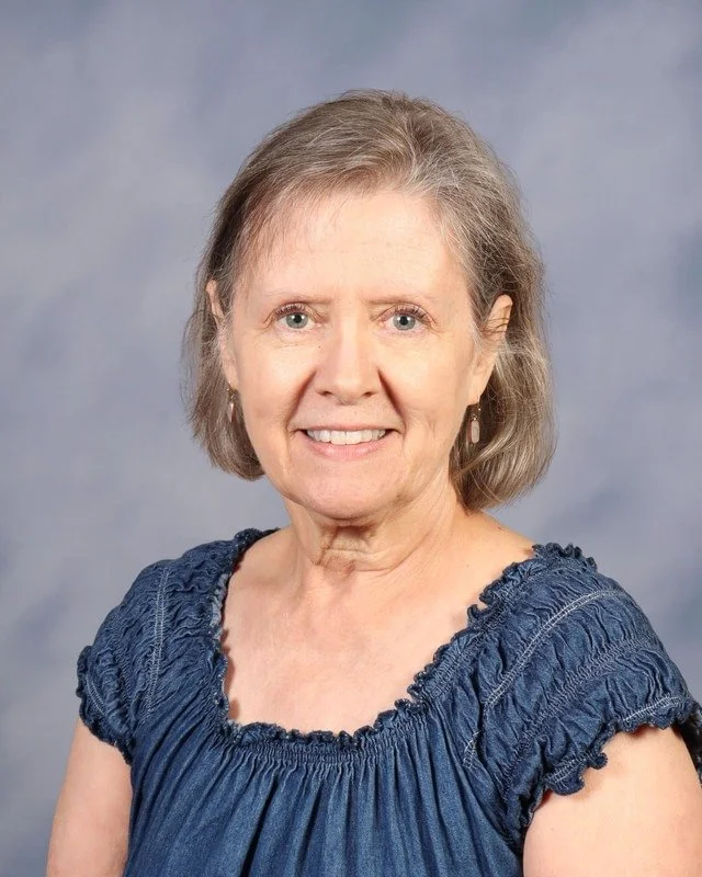 A smiling elderly woman with short gray hair, wearing a white t-shirt and a cross necklace, standing outdoors with blurred green trees in the background.