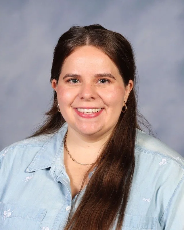 A smiling woman with long brown hair wearing a white blouse, standing outdoors in a park with trees in the background.