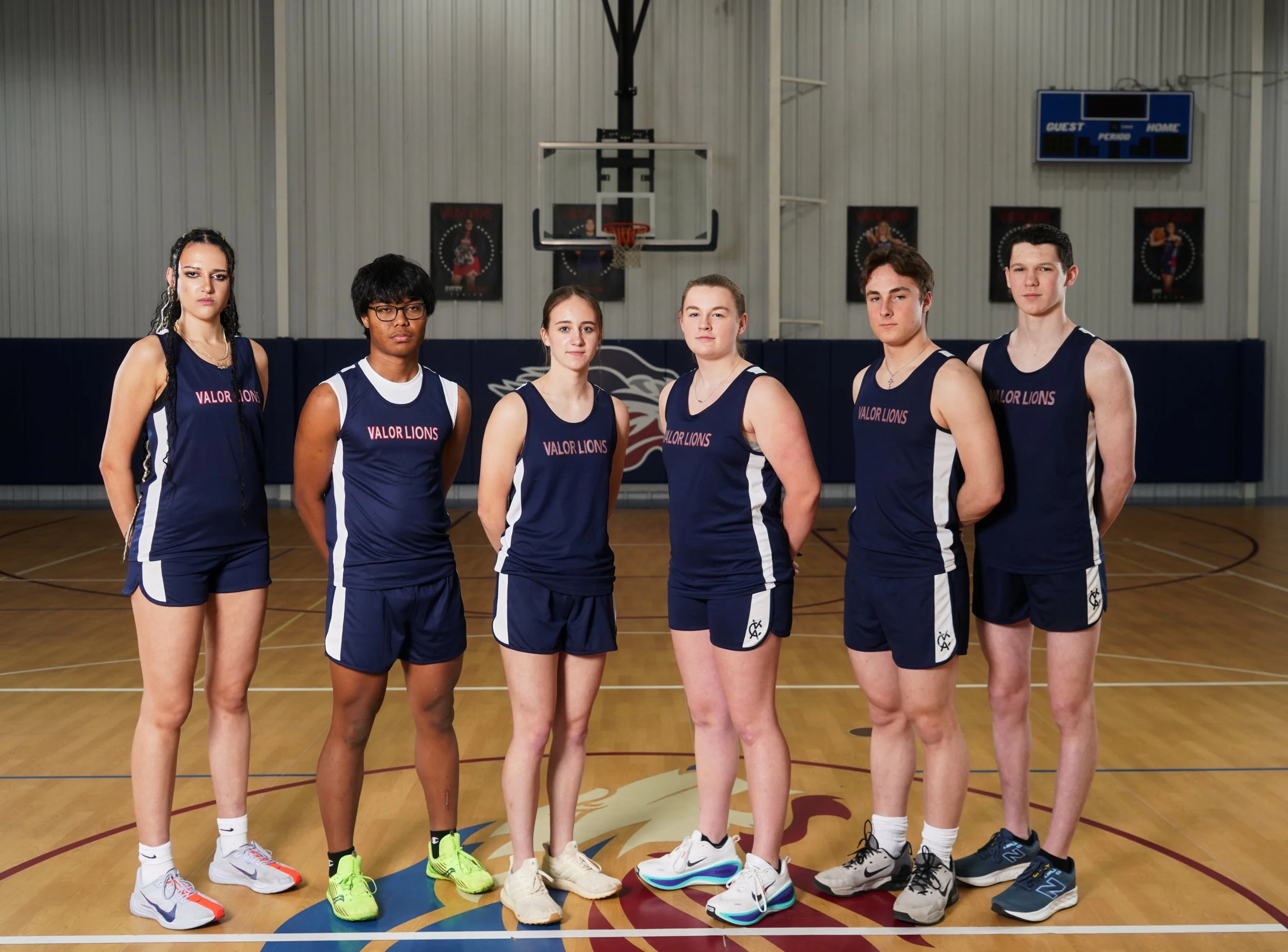 A girls' basketball team and coach posing on a gymnasium basketball court. The team members are wearing navy uniforms with IVALOR LIONS written on them. They are standing and kneeling in front of a basketball hoop.