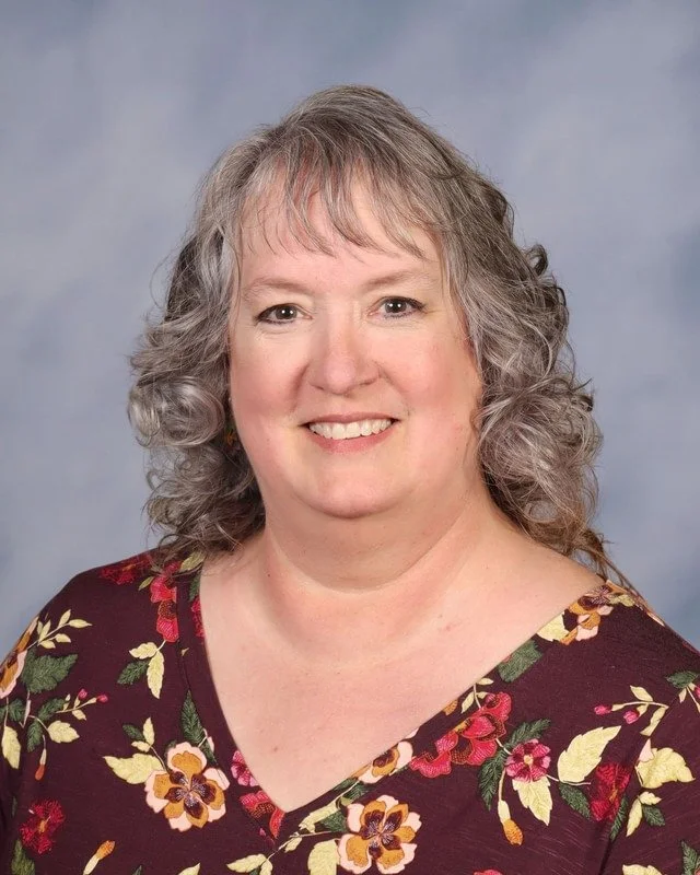 A smiling woman with gray hair wearing a colorful, knitted shawl with blue, green, gray, black, and tan colors, standing outdoors in a park with trees in the background.