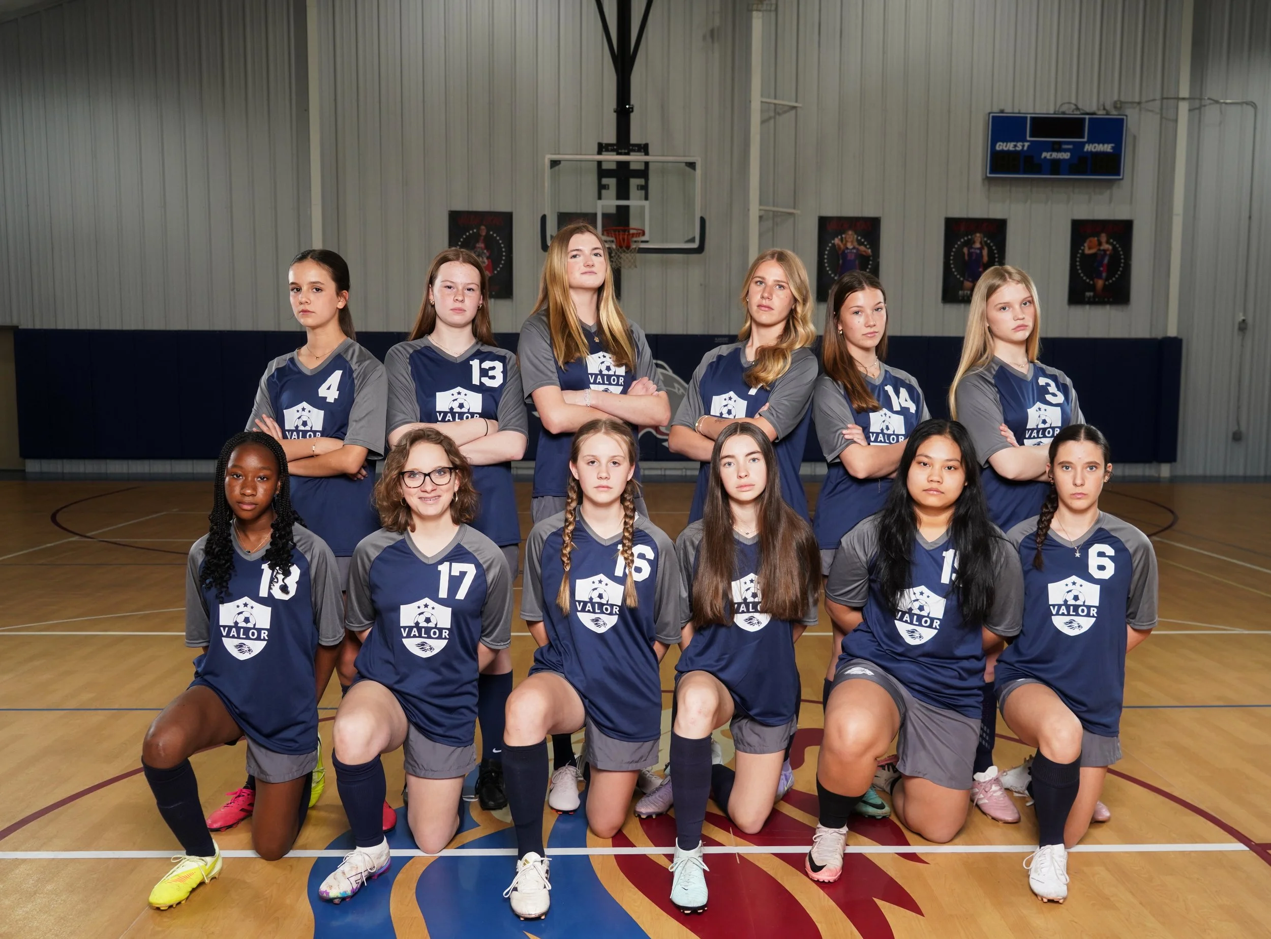A girls' volleyball team with coach and assistant coach in a gymnasium, wearing blue and gray uniforms with the team logo, posing for a team photo with two volleyballs, in front of a Valor Lions logo on the floor.