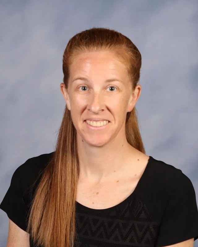 A woman with long red hair wearing a light gray short-sleeve top standing outside in a park with green grass and trees in the background.