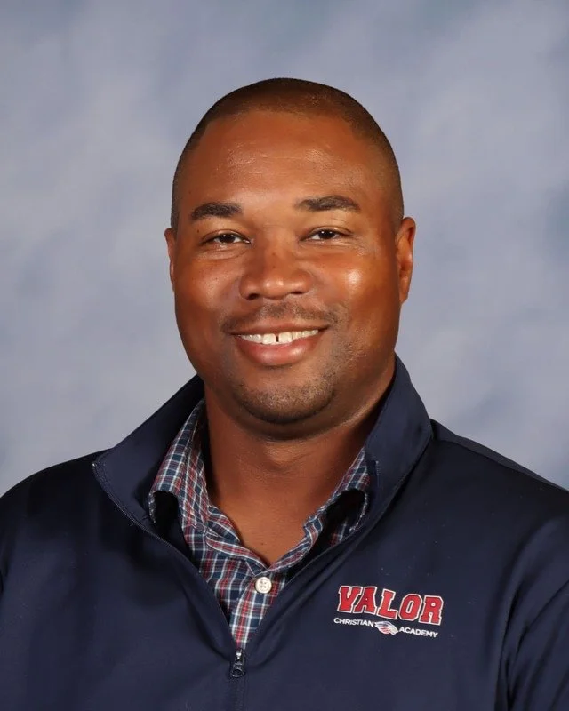 A man standing outdoors on a grassy area, smiling and wearing a white polo shirt, with trees and a fence in the background.