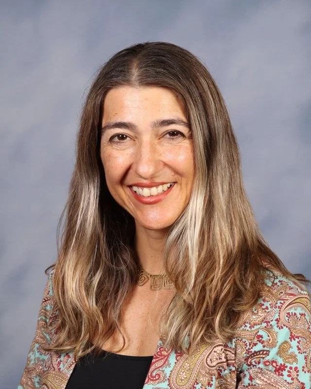 A smiling woman with wavy blonde hair in a white collared shirt and dark blazer outdoors in a green park.