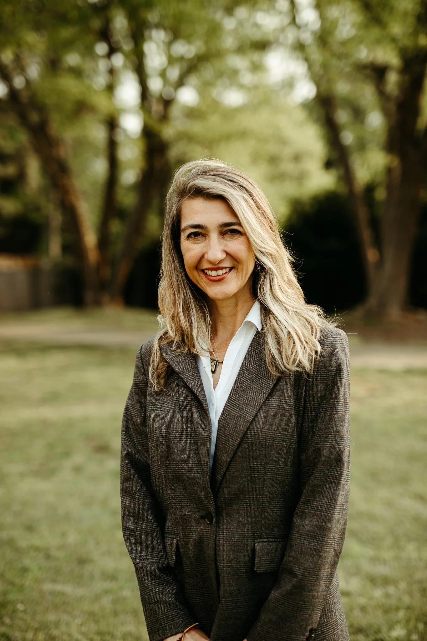A woman with long blonde hair, wearing a brown plaid blazer over a white shirt, smiling outdoors in a grassy park with trees in the background.