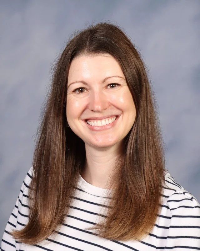 A woman with long brown hair smiling and standing outdoors in a park with trees in the background.