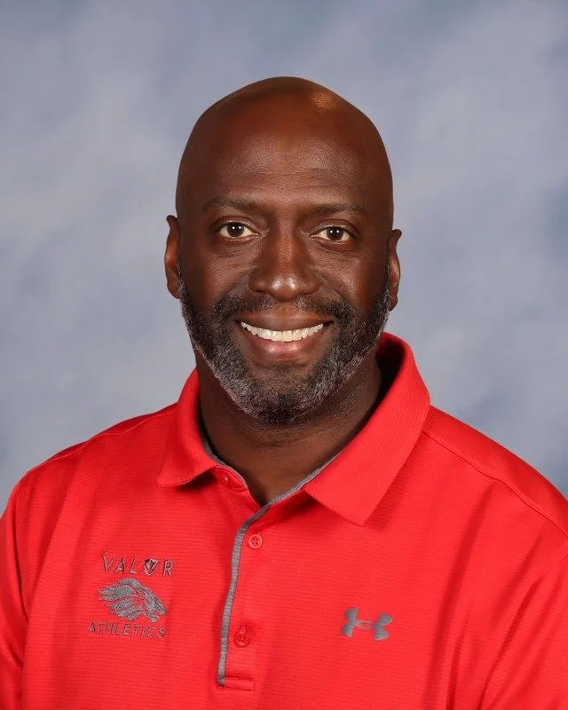 Smiling man outdoors wearing a blue and gray sports jacket with 'Valor Athletics' logo, with trees in the background.
