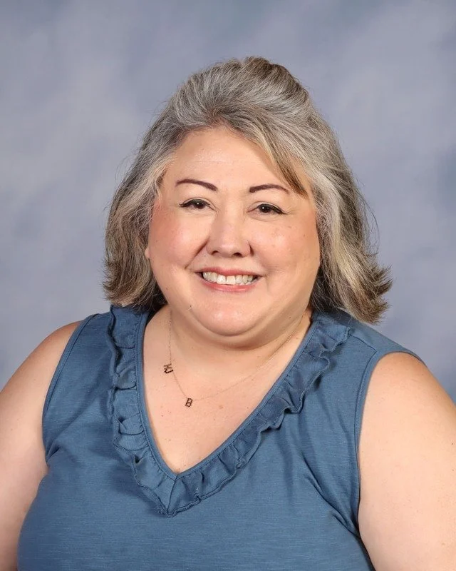 A middle-aged woman with gray hair smiling outdoors in a park with trees in the background, wearing a black jacket over a white top.