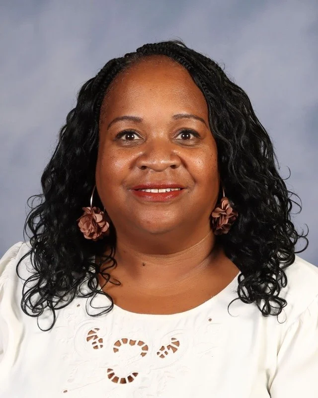 A woman with curly hair wearing a red blouse standing outdoors in a park with trees in the background.