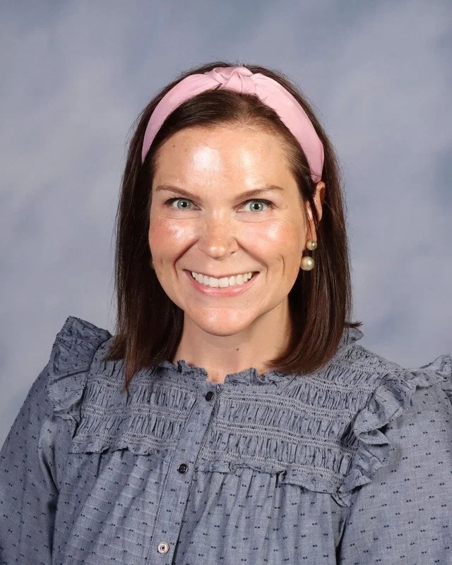 A woman smiling outdoors wearing a red dress with ruffled sleeves and a blue headband, standing on grass with trees in the background.