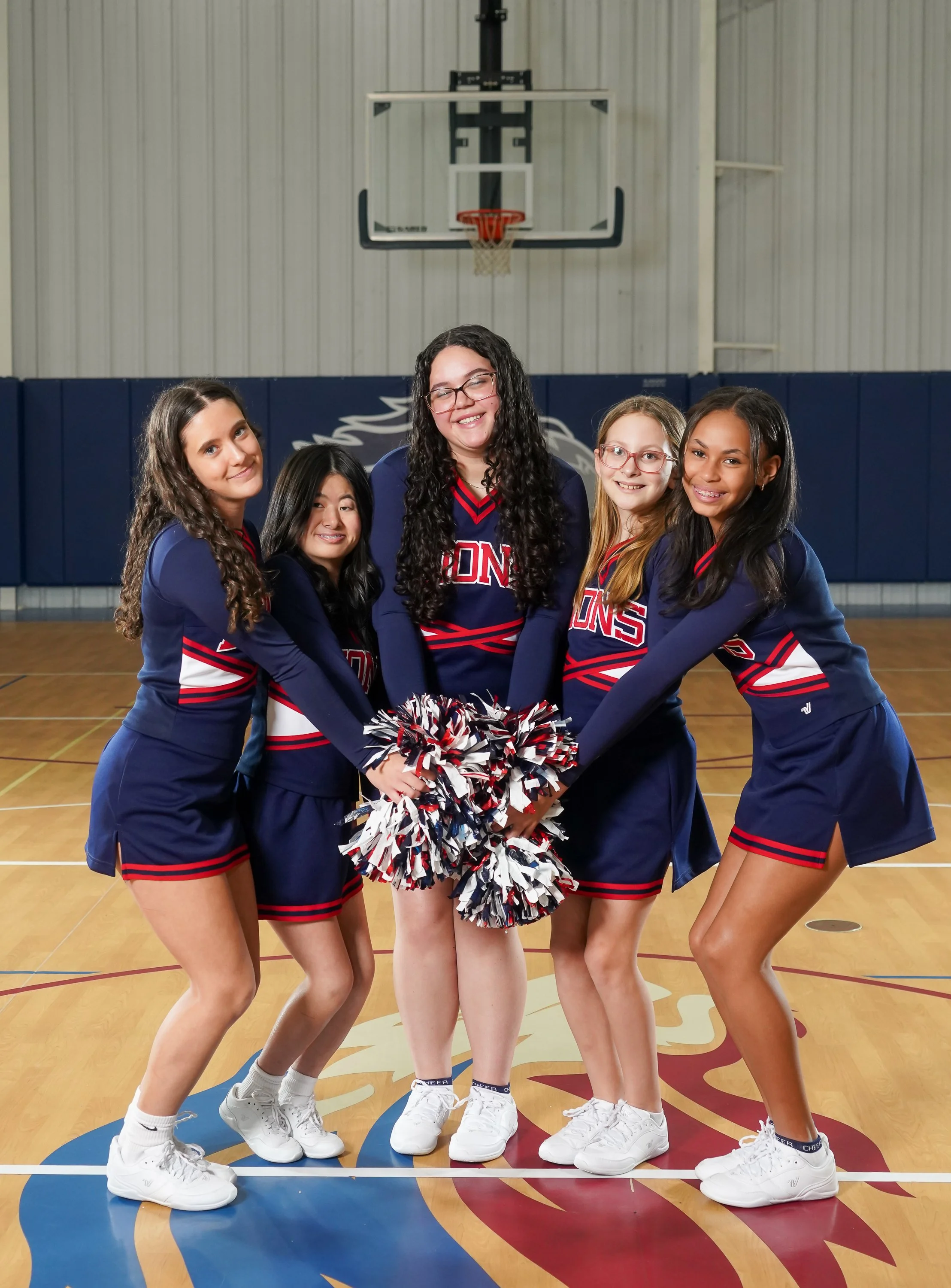 Eight female cheerleaders in navy blue uniforms with red and white accents, holding pom-poms, standing in a gymnasium with a basketball hoop and banners in the background.