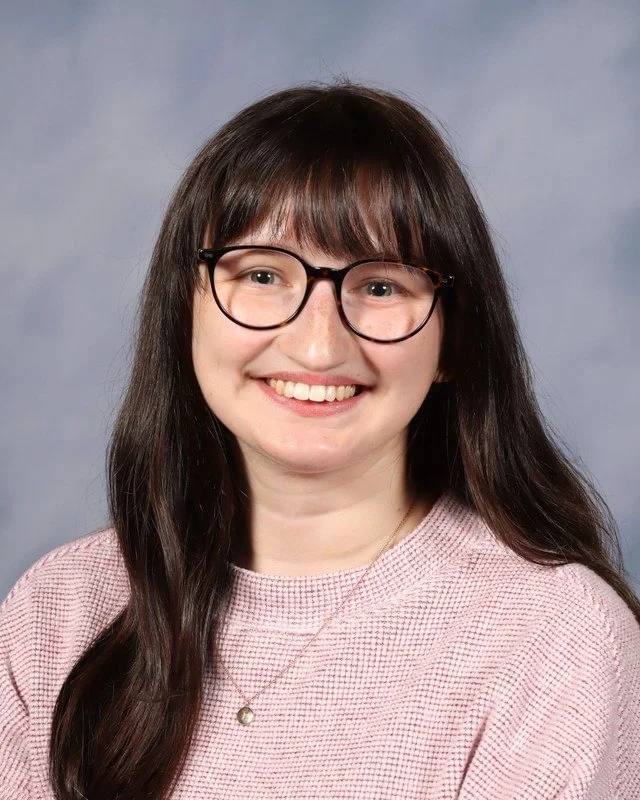 A young woman with long brown hair, wearing a navy blue t-shirt, smiles standing outdoors in a park with green grass and trees in the background.