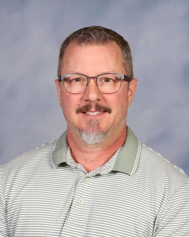 A man with glasses and a trimmed beard, wearing a navy blue polo shirt with 'Valor Christian Academy' embroidered on the left chest, standing outdoors with trees in the background.