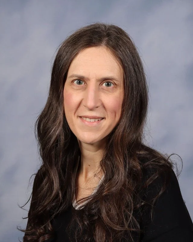 A woman with long brown hair in a blue blouse standing outdoors in a park with trees in the background.