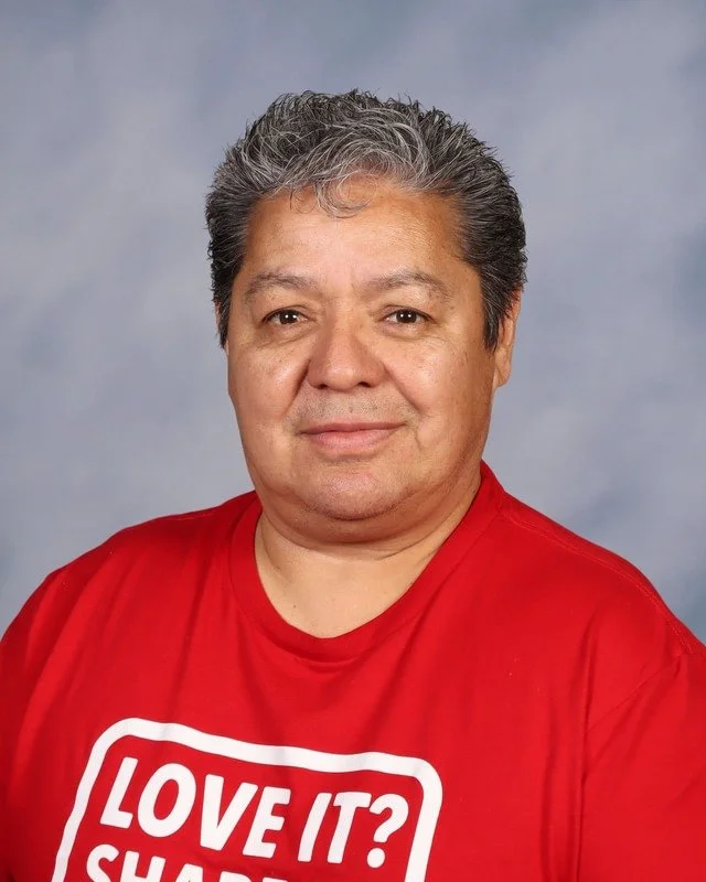 A smiling man with gray hair wearing a navy blue t-shirt with 'VALOR Christian Academy' written on it, standing outdoors with trees and grass in the background.