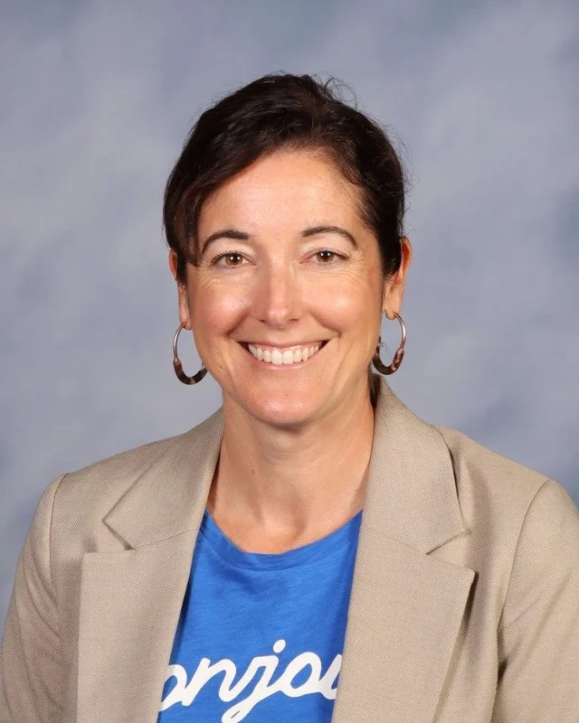 A woman with shoulder-length dark hair smiling outdoors in a park with green trees and grass, wearing a white shirt, blue blazer, and brown belt.