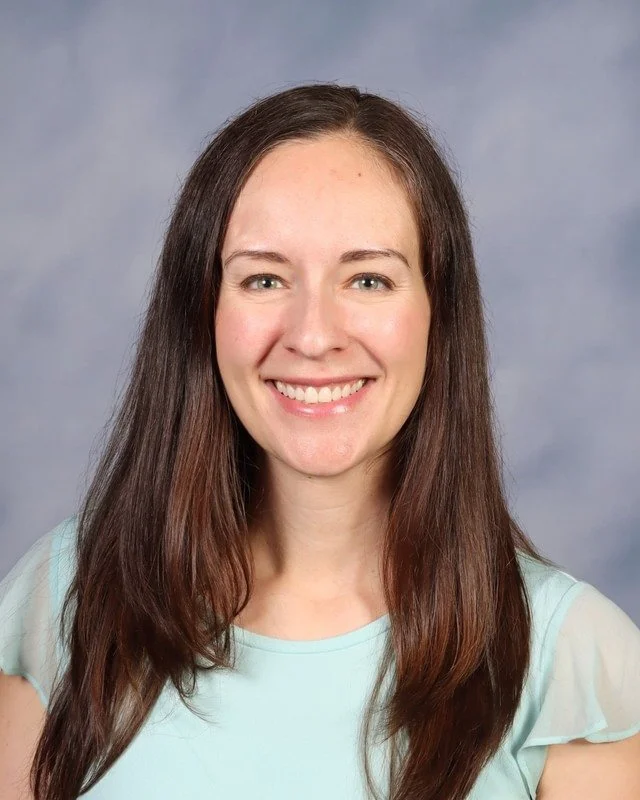 A woman with long brown hair wearing a black and white striped shirt smiling outdoors in a park with trees and grass in the background.