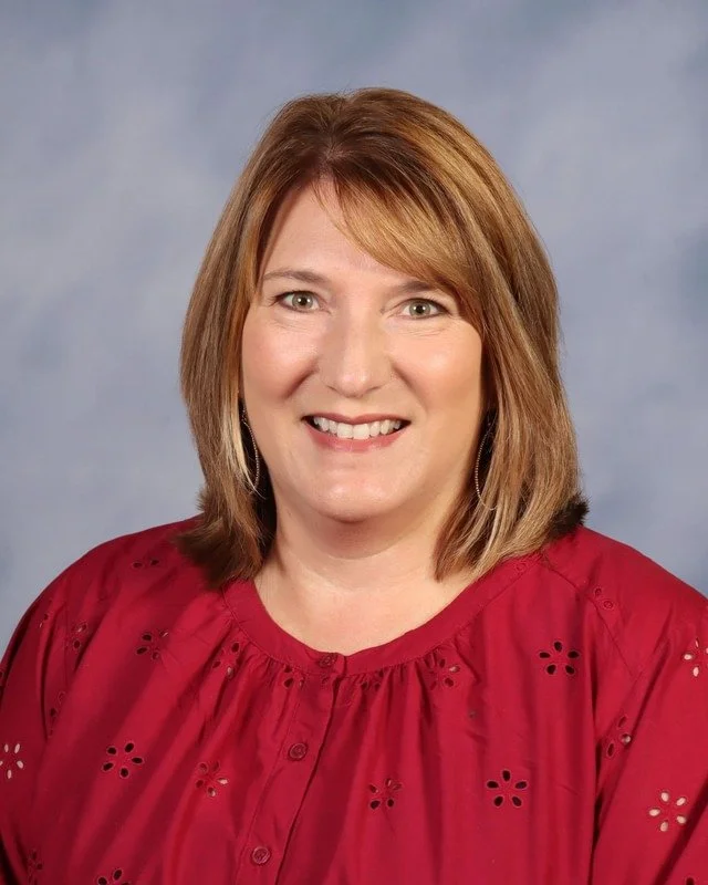 A woman with shoulder-length light brown hair, smiling, standing outdoors with greenery in the background, wearing a dark blue blouse and white pants.