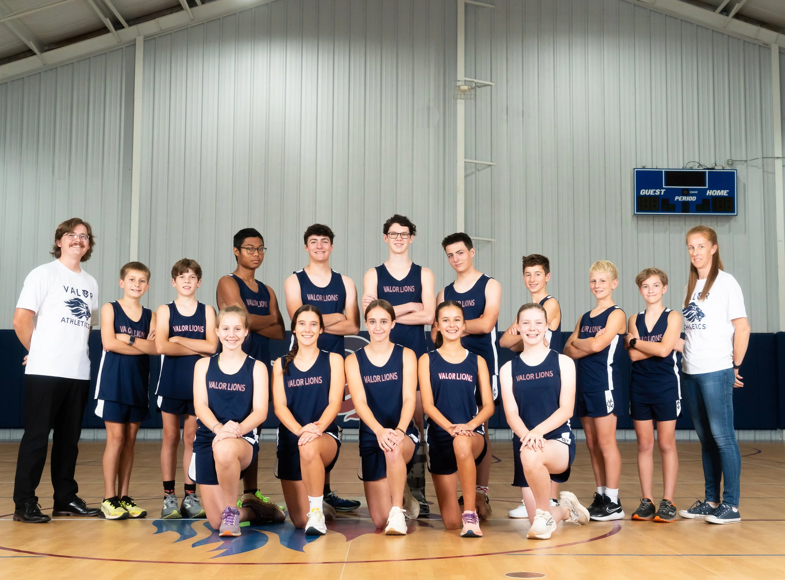 A girls' basketball team posing for a photo in a gymnasium. The team, called Valor Lions, consists of six young women in navy blue sports uniforms and a coach or team staff member in a gray hoodie and jeans. They are standing on a basketball court with a team logo on the floor and a scoreboard in the background.