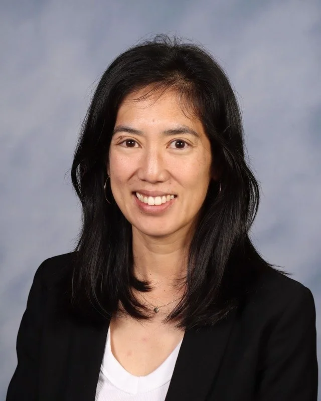 A woman with long black hair, wearing a dark blue top and hoop earrings, stands outdoors in a park with grassy area and large trees in the background, smiling at the camera.