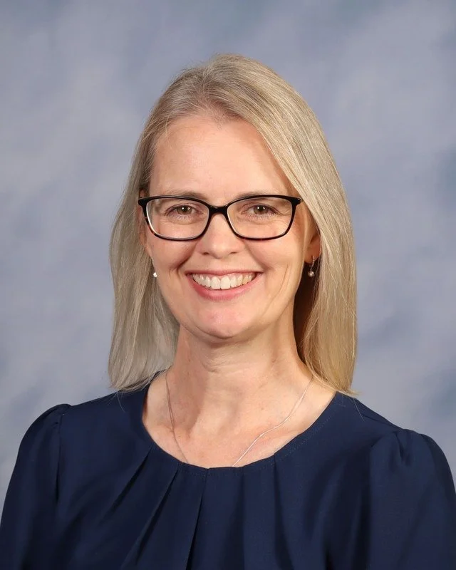 A smiling woman with blonde hair, glasses, and a blue dress standing outdoors with trees in the background.