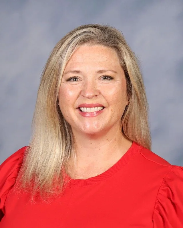 A woman smiling outdoors in a pink dress with ruffled sleeves, standing in a park with trees in the background.