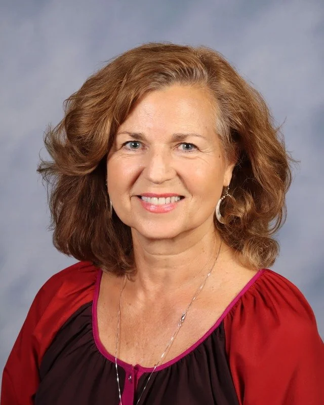 A middle-aged woman with wavy, shoulder-length hair, smiling, wearing a white shirt and a long necklace, standing outdoors in a park with trees in the background.