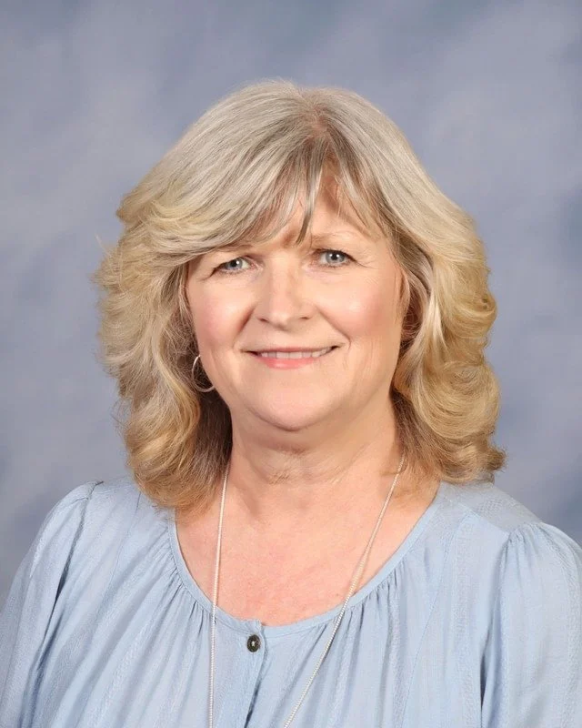 A middle-aged woman with blonde hair smiling outdoors in a park with green trees in the background, wearing a maroon top and layered necklaces.