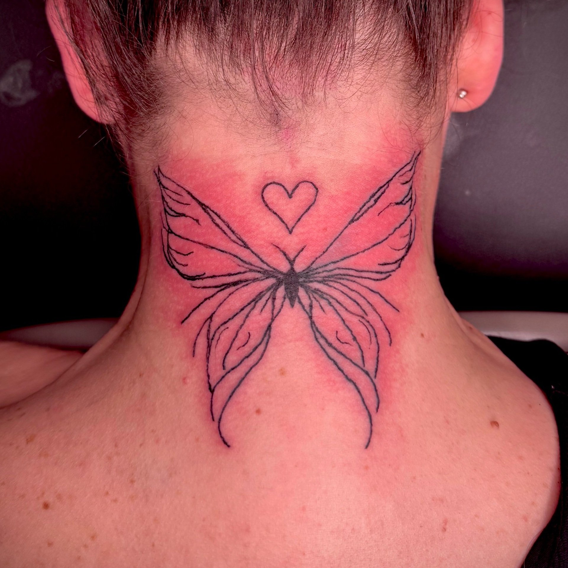 Tattoo of a butterfly with a heart above it on the back of a person's neck.