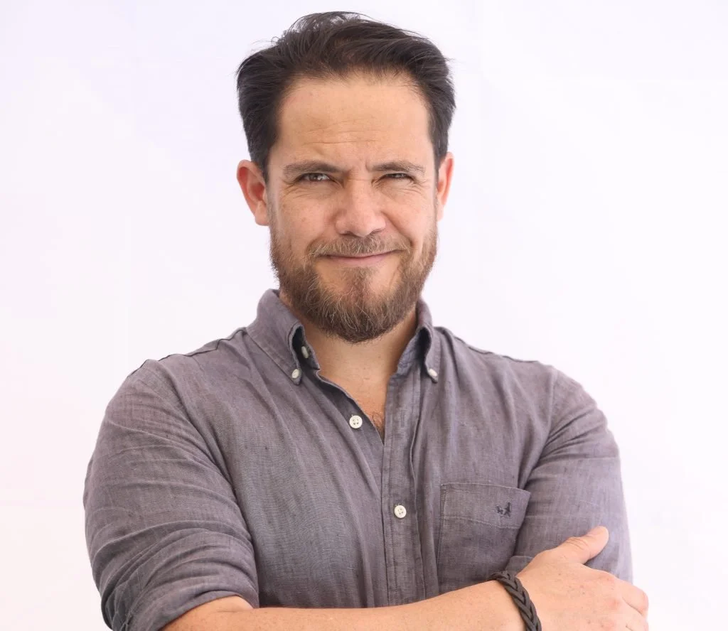 Hombre con barba y camisa gris sonriendo ligeramente, brazos cruzados, fondo blanco.